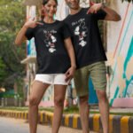 A South Asian couple wearing Hinglish t-shirts. The girl's shirt reads "He's Mine" and the boy's shirt reads "She's Mine."